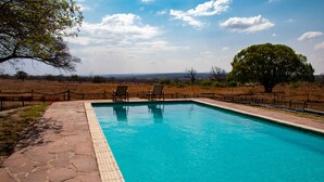 A rectangular pool with a tiled edge, surrounded by a stone walkway. There are two lounge chairs facing the pool, and a wooden fence enclosing the area. - Waterberg Cottages (Vaalwater)