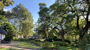 A well-maintained garden with a variety of plants and trees, a stone pathway, and a rustic house with a thatched roof. - Waterberg Cottages (Vaalwater)