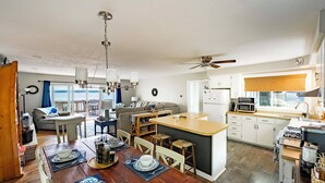 A modern kitchen with a central island, white cabinetry, and a dining area with a wooden table set for four. - Waterfront Deck + Dock: Torch Lake Home! (Rapid City)