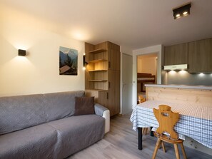 A cozy room with a sofa, a dining table with a checkered tablecloth, wooden shelves, and a kitchen area with cabinets. - Cozy studio with pets allowed, near slopes and shops (La Clusaz)