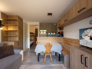 Une cuisine moderne avec des meubles en bois, une table à manger recouverte d’une nappe à carreaux et un téléviseur fixé au mur.