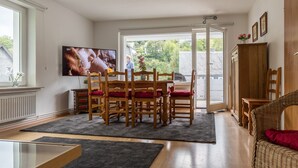 Comedor con mesa y sillas de madera, televisor en la pared y vista a zonas verdes al exterior.