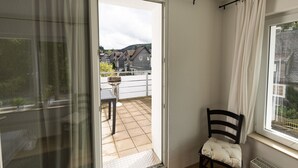Un balcón con una silla y una parrilla, una puerta que da al exterior y vistas a casas.