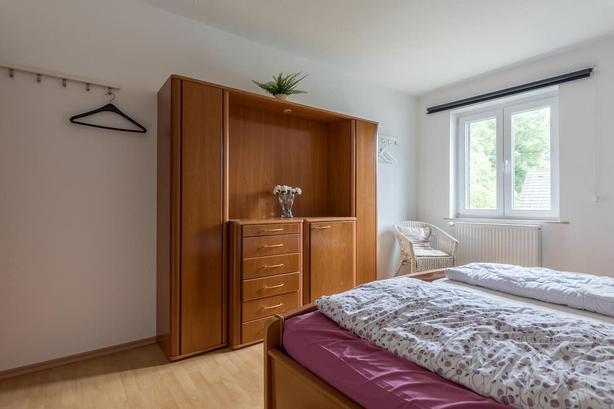 Ein Schlafzimmer mit einem Holzschrank, einem Bett mit gemusterten Textilien, einem Sessel und einem Fenster mit Blick auf Grünflächen.
