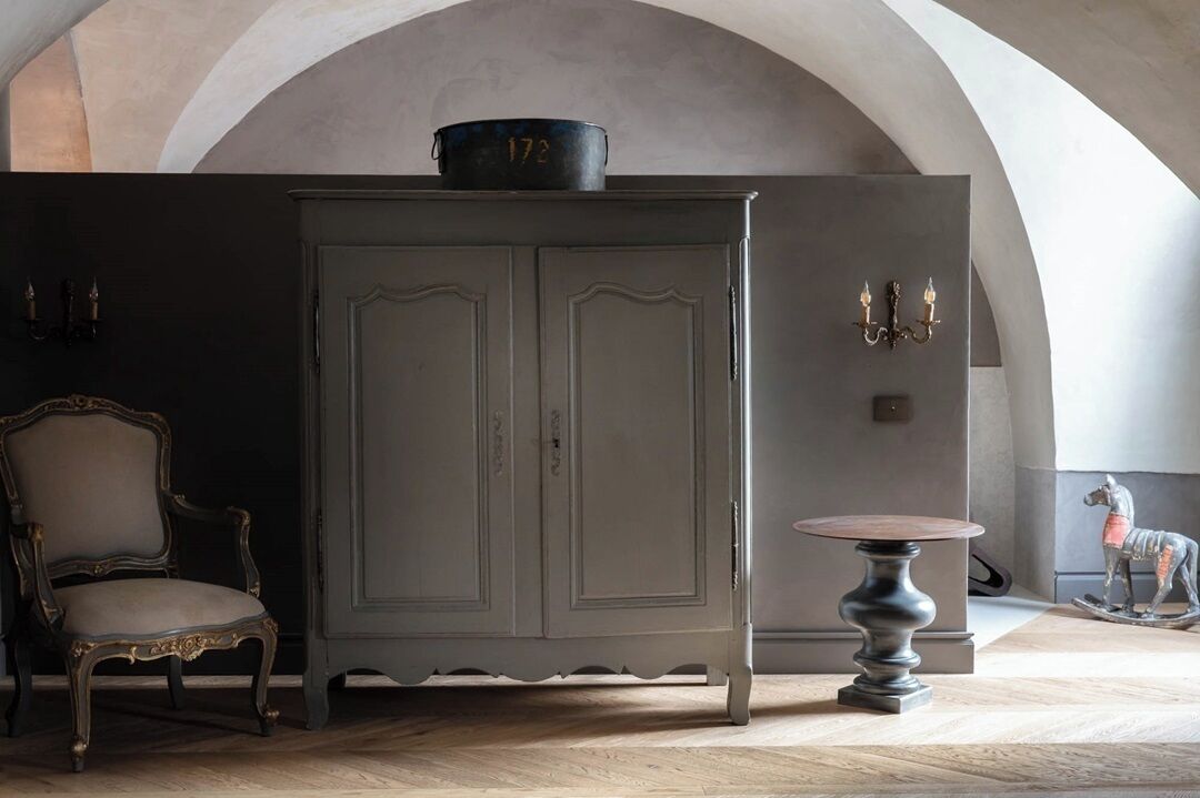 A room with a dark grey arched ceiling, a vintage-style cabinet with a number '172' on top, a classic chair, a round table, and a small horse statue.