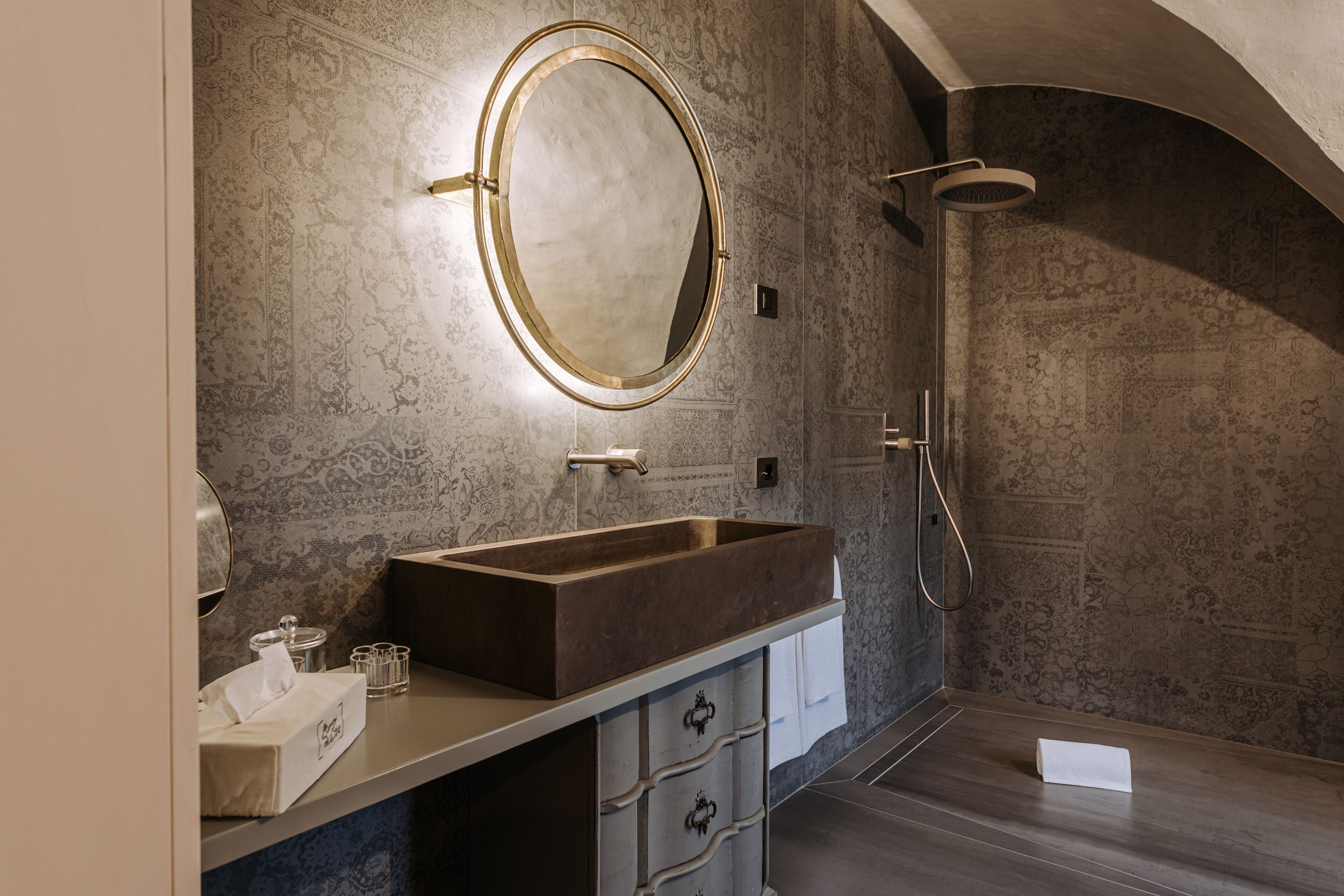 Un baño con un espejo ovalado grande, un tocador de madera, ducha y paredes con diseño.