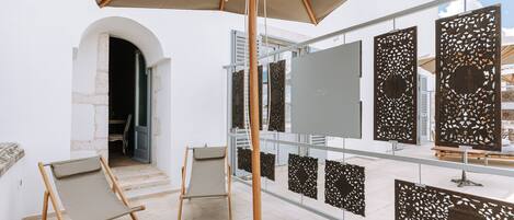 An art gallery with a glass wall displaying various artworks. There are two wooden chairs and a large umbrella providing shade.