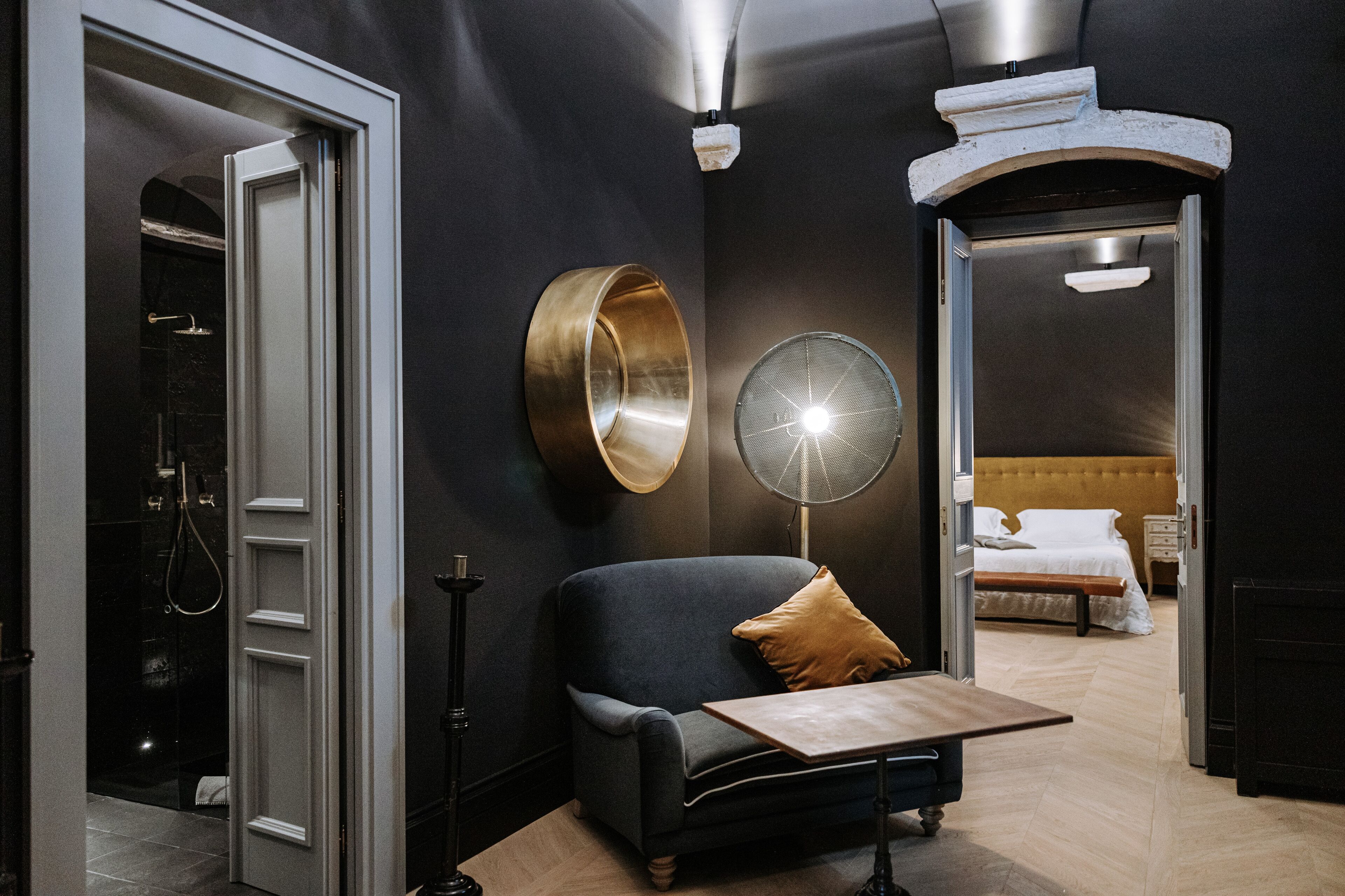 Una camera d'albergo moderna con un divano scuro, un tavolino, una lampada da terra rotonda e un letto con lenzuola bianche.
