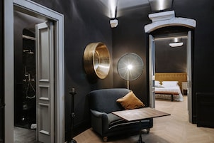 Una camera d'albergo moderna con un divano scuro, un tavolino, una lampada da terra rotonda e un letto con lenzuola bianche.