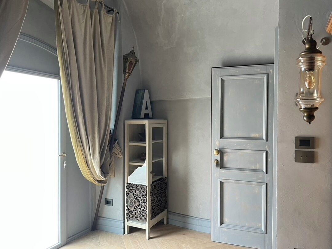 A room with a light blue door, a small white shelf with a letter 'A' on it, a hanging lantern, and a window with curtains.
