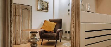A room with a wooden floor, a brown armchair with a mustard-colored pillow, a small round table, and a wall-mounted shelf with glasses.