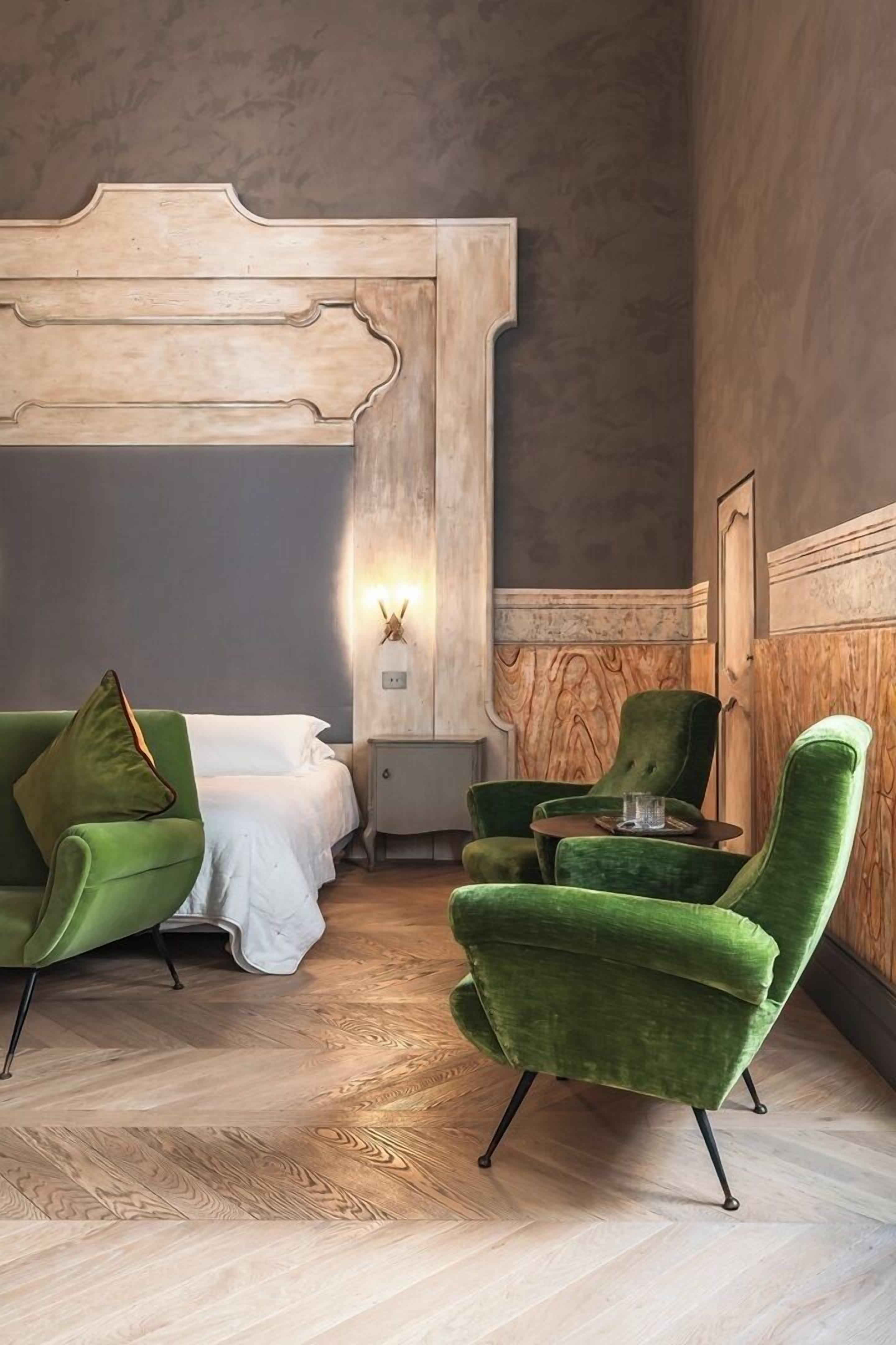Una habitación de hotel con cama, sillones verdes, una mesita y una lámpara fijada a la pared.
