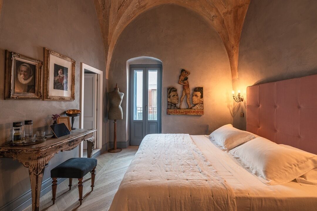 A bedroom with a large bed, a desk, and a chair. There are framed pictures on the wall and a window with a view.