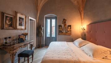 A bedroom with a large bed, a desk, and a chair. There are framed pictures on the wall and a window with a view.