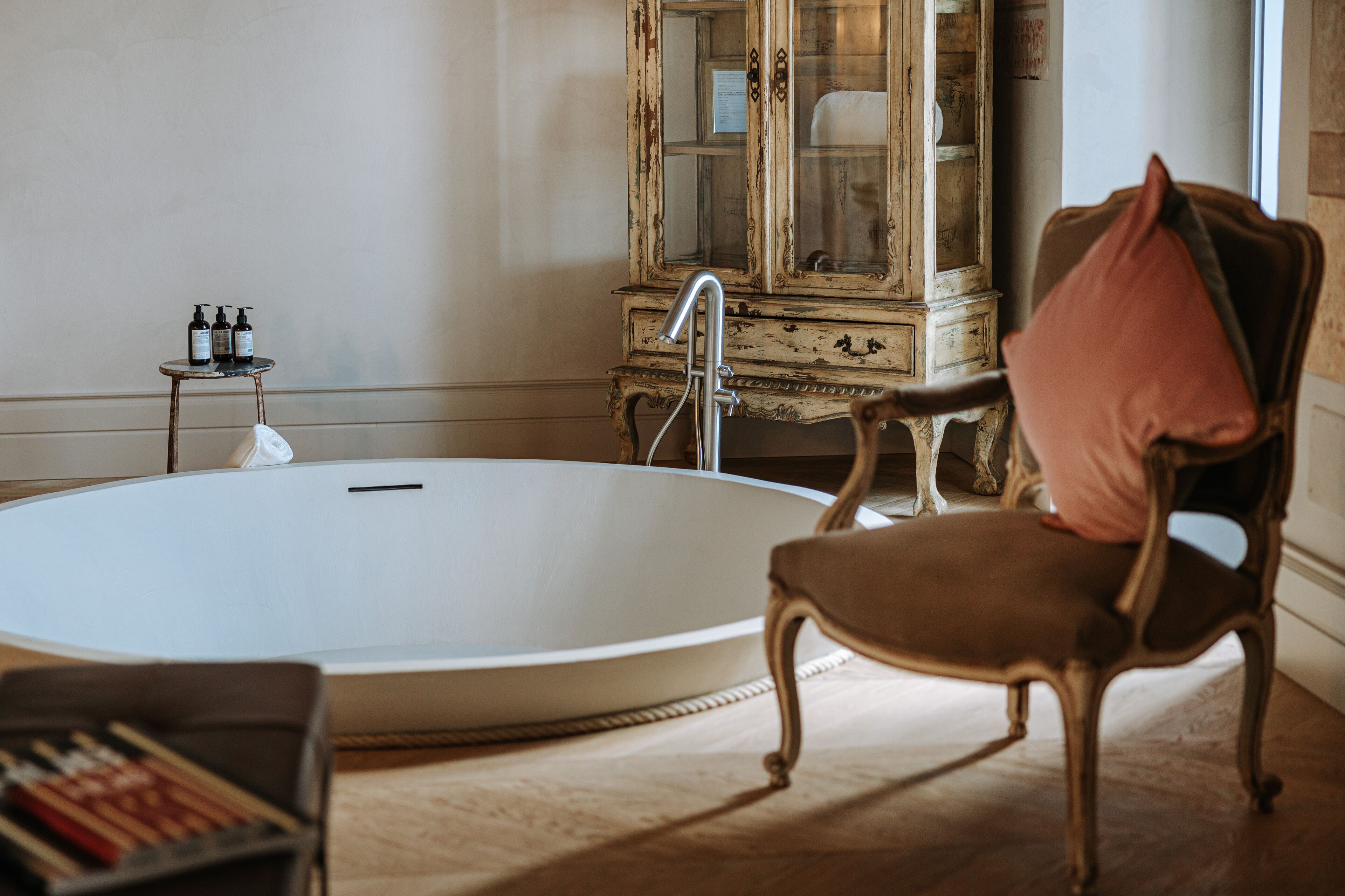 Una habitación con una bañera ovalada grande, un aparador de madera antiguo y una silla con cojín.