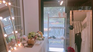 A room with a vanity table, mirror, and lights. There is a window with a view of greenery outside. - Wedding House (Seogwipo)