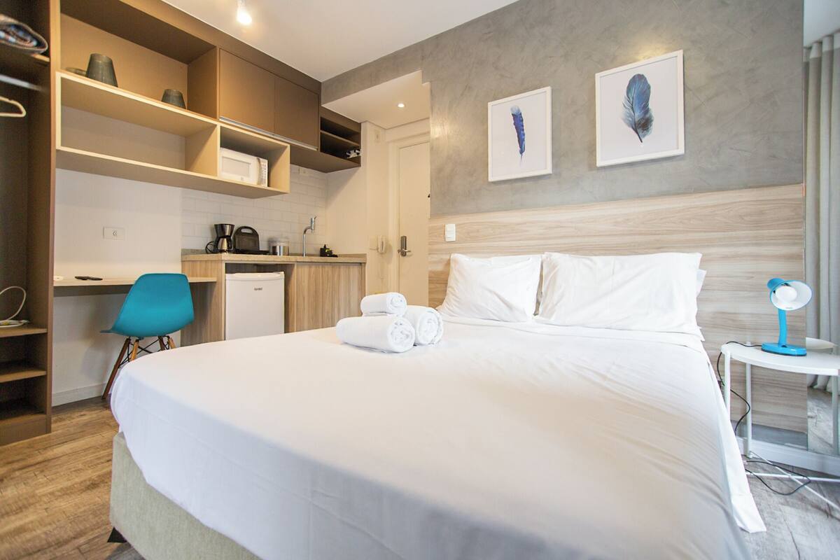 Una habitación de hotel con cama, escritorio, silla, una mesita con lámpara y cuadros en la pared.
