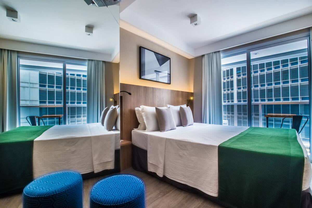Habitación de hotel con dos camas, un ventanal grande y vistas a la ciudad.