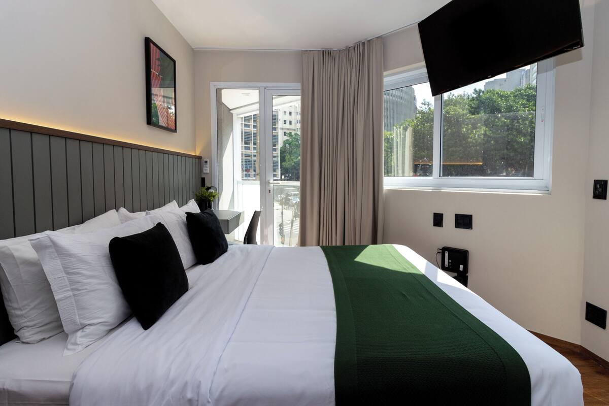 Habitación de hotel con una cama grande, un televisor de pantalla plana montado en la pared, una ventana con vistas a la ciudad y una puerta que da a un balcón.