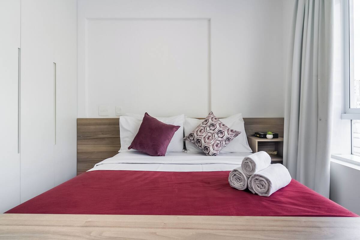 Una cama bien hecha con una colcha roja, dos almohadas y toallas enrolladas sobre un piso de madera.