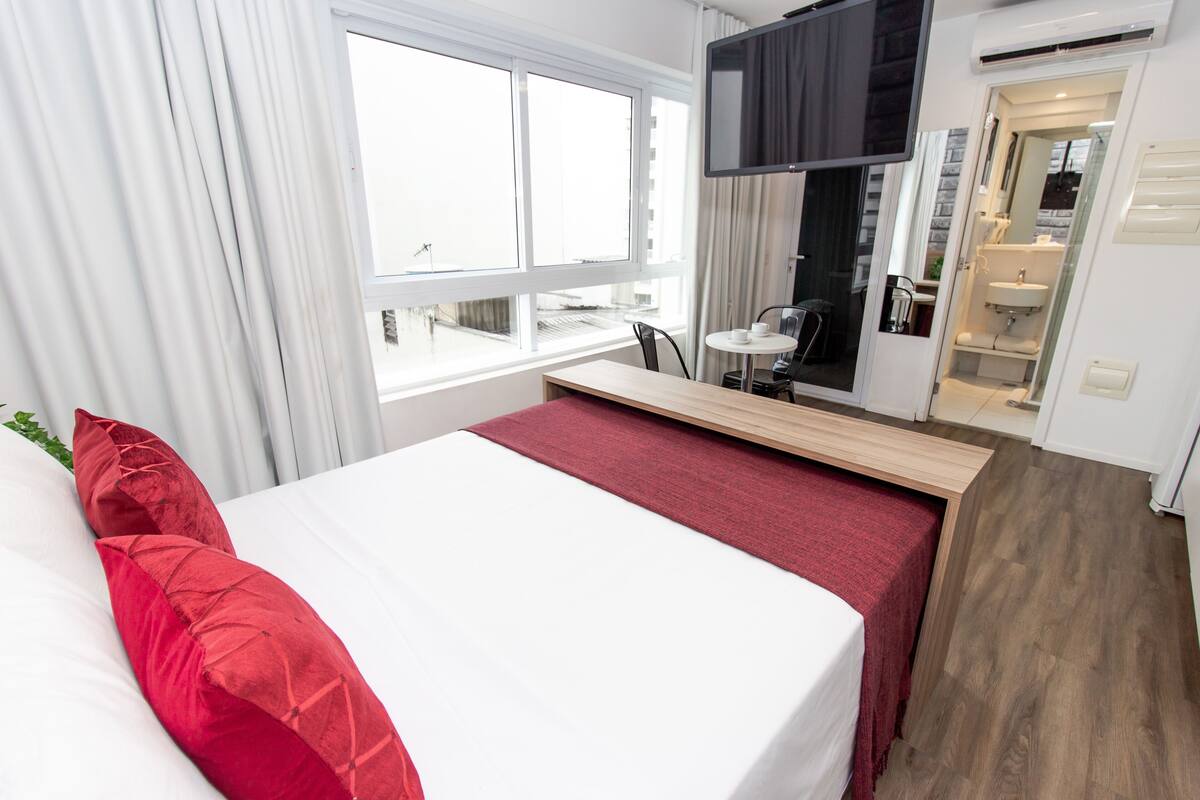 Habitación de hotel con cama, televisor, una mesita y baño visible a través de una puerta de cristal.