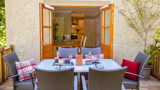 Patio aperto con tavolo apparecchiato per due, con sedie in vimini e una bottiglia di champagne.