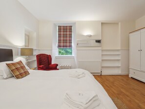 Un dormitorio con una cama, un sillón rojo, una ventana con cortina escocesa, un radiador y un armario.