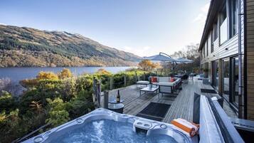 Relax on the deck with a hot tub, sofa, and chairs, overlooking a serene lake and majestic mountains.