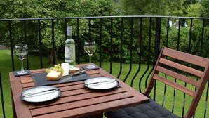 Una mesa de madera dispuesta con una botella, queso, uvas y cubiertos en un balcón con barandilla de metal y abundante vegetación al fondo.