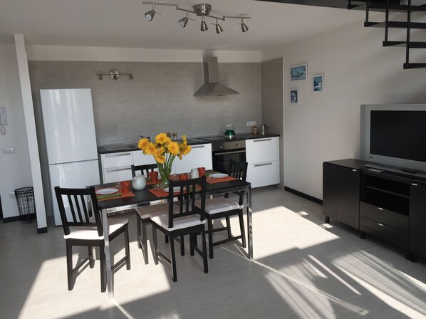 A modern kitchen with a dining area, featuring a central island with chairs, a refrigerator, a stove, and a television on a cabinet.