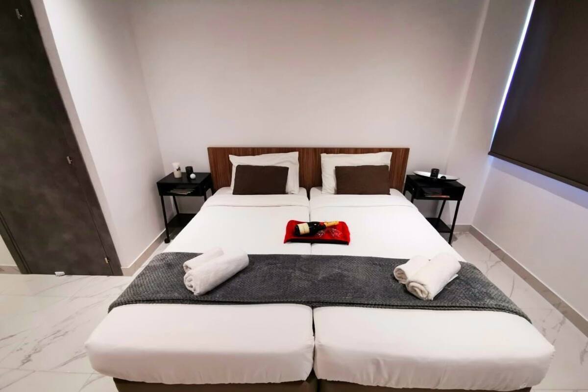 Uma cama de casal com um cobertor cinza e toalhas, duas mesas de cabeceira com abajures, e uma porta de madeira escura.