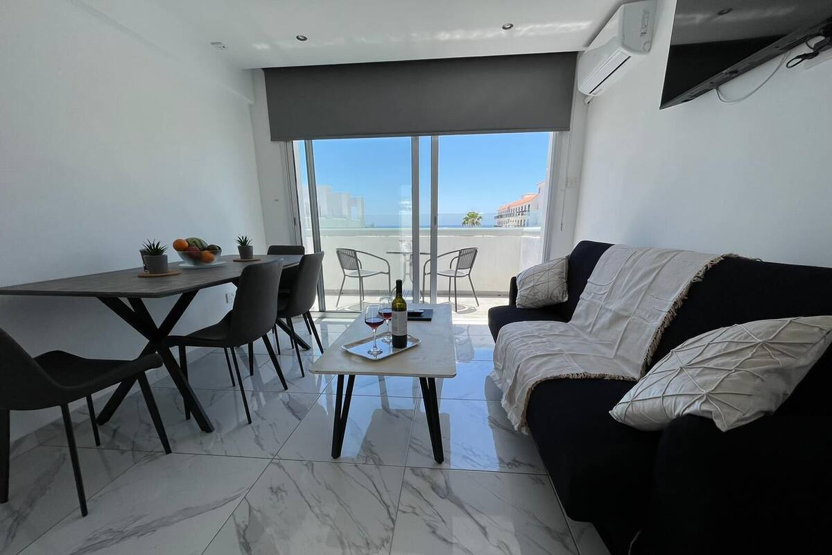 Uma sala de estar moderna com área de jantar, um sofá preto e uma grande janela com vista para o mar.