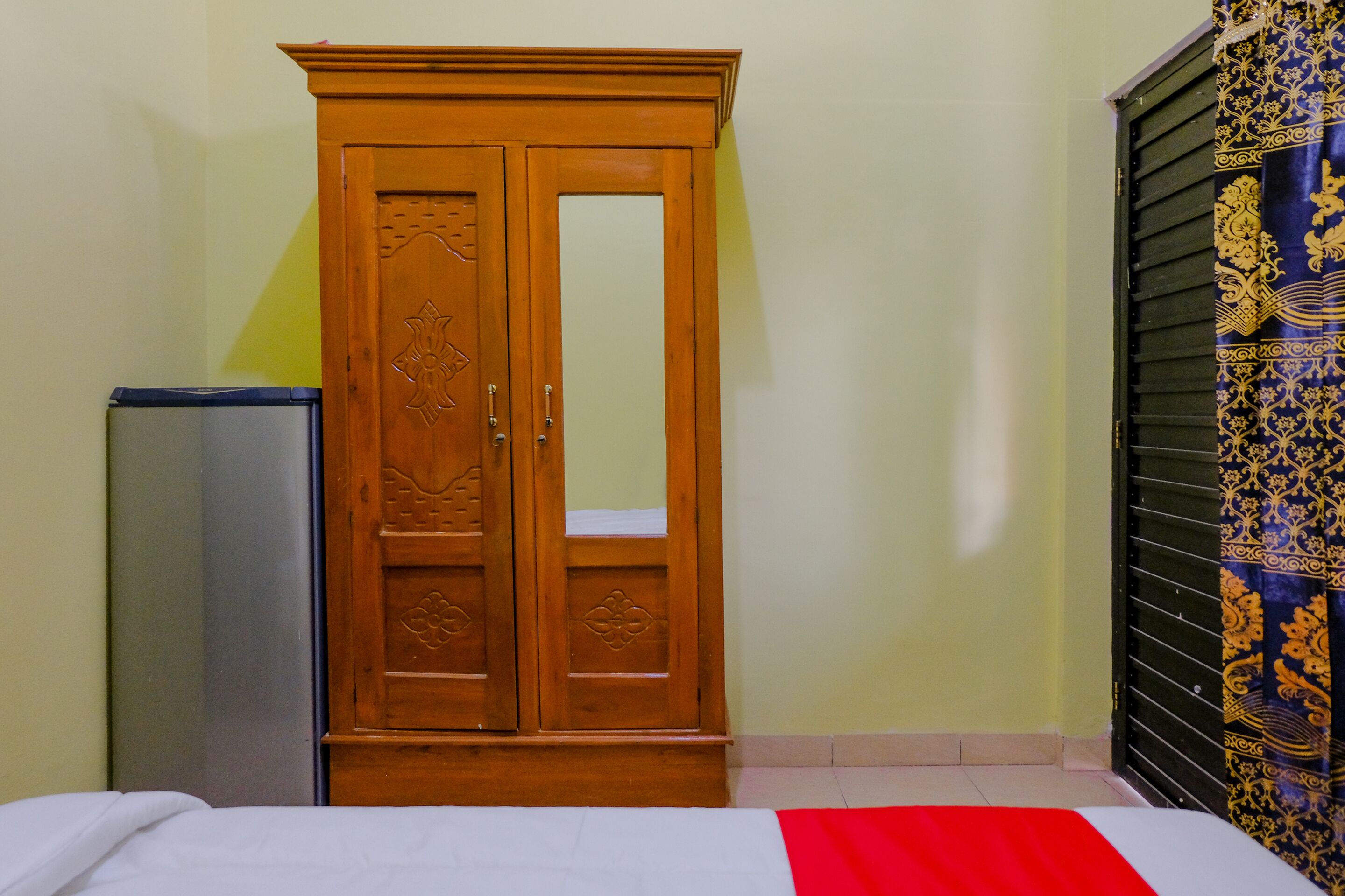 Quarto com guarda-roupa de madeira, cama com colcha branca e vermelha, e cortina estampada na janela.
