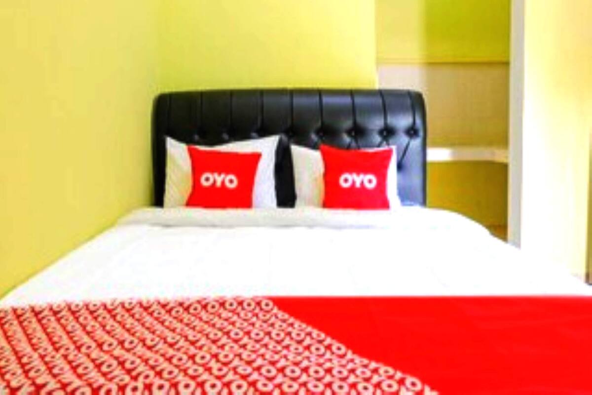 Un lit avec une tête de lit noire, deux oreillers rouges ornés du mot « OYO » et une couvre-lit blanche et rouge à motifs.