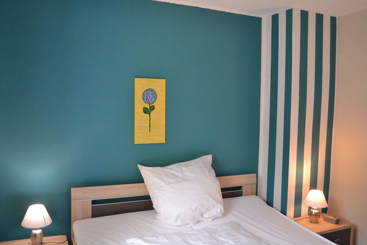 Une chambre avec des murs de couleur turquoise, une tête de lit en bois, un oreiller blanc, une œuvre d’art jaune représentant une fleur et un mur à rayures.