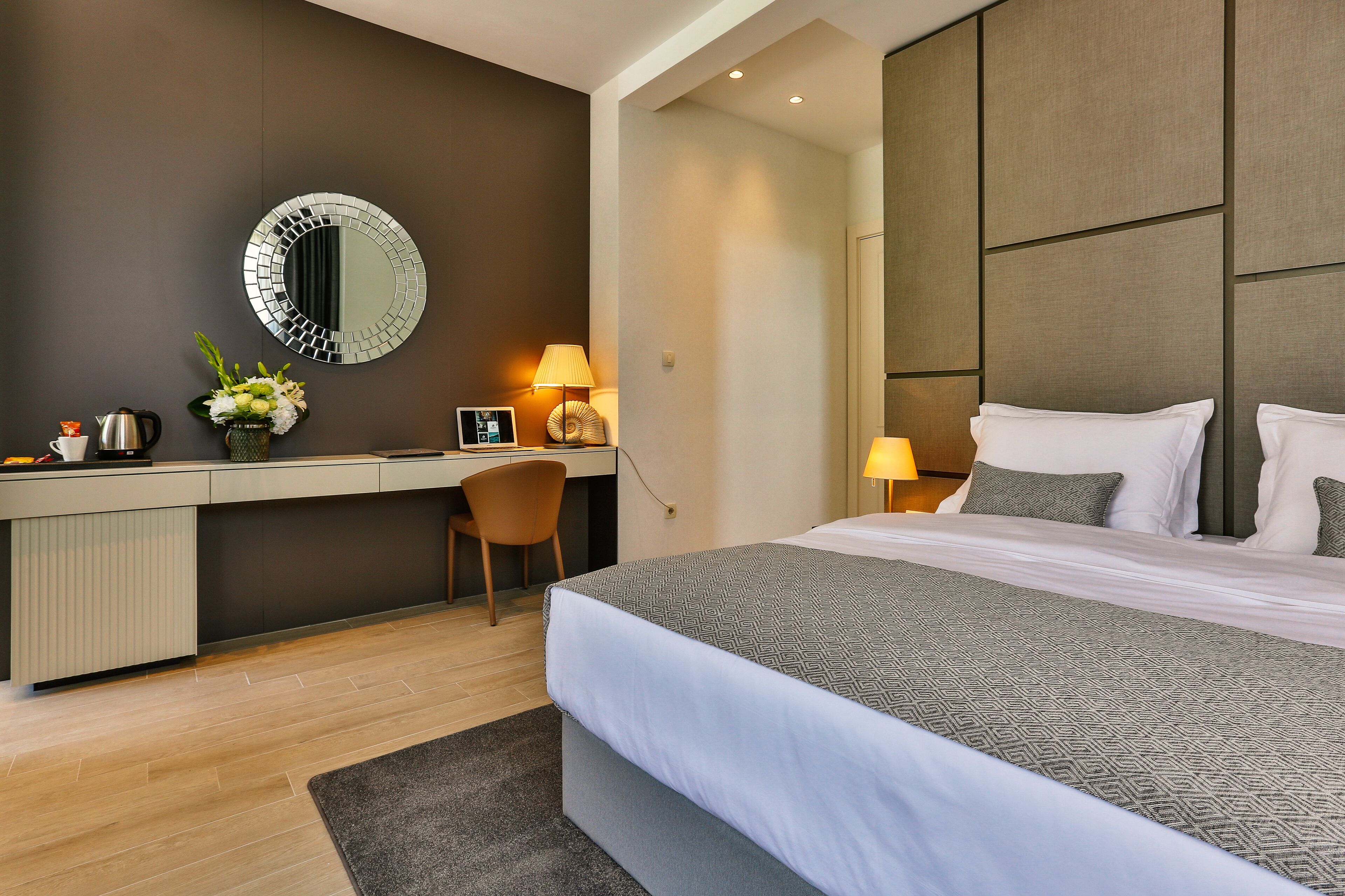 A modern hotel room with a large bed, a desk, a chair, a round mirror, and two bedside lamps.