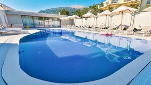 Piscina con sillas de descanso y sombrillas, rodeada por un edificio con vistas a montañas.