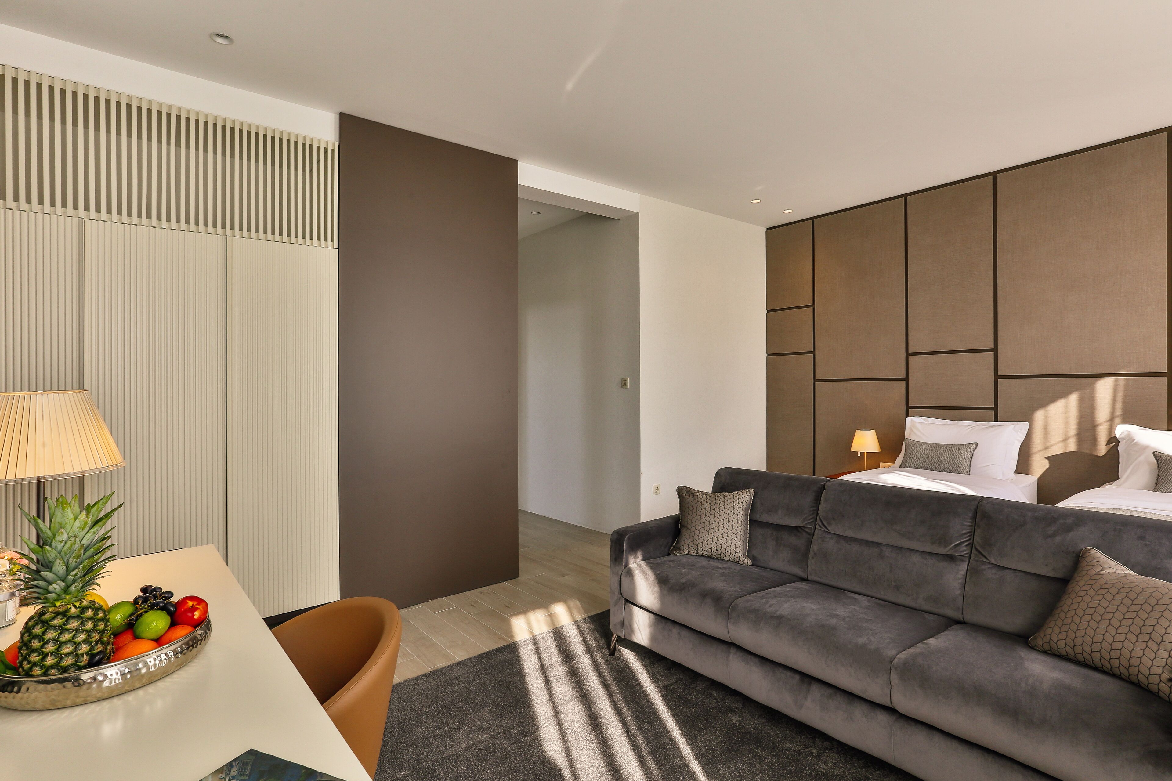 Une chambre d’hôtel moderne avec un canapé gris, une table à manger avec des fruits, une lampe et un lit doté d’une tête de lit.
