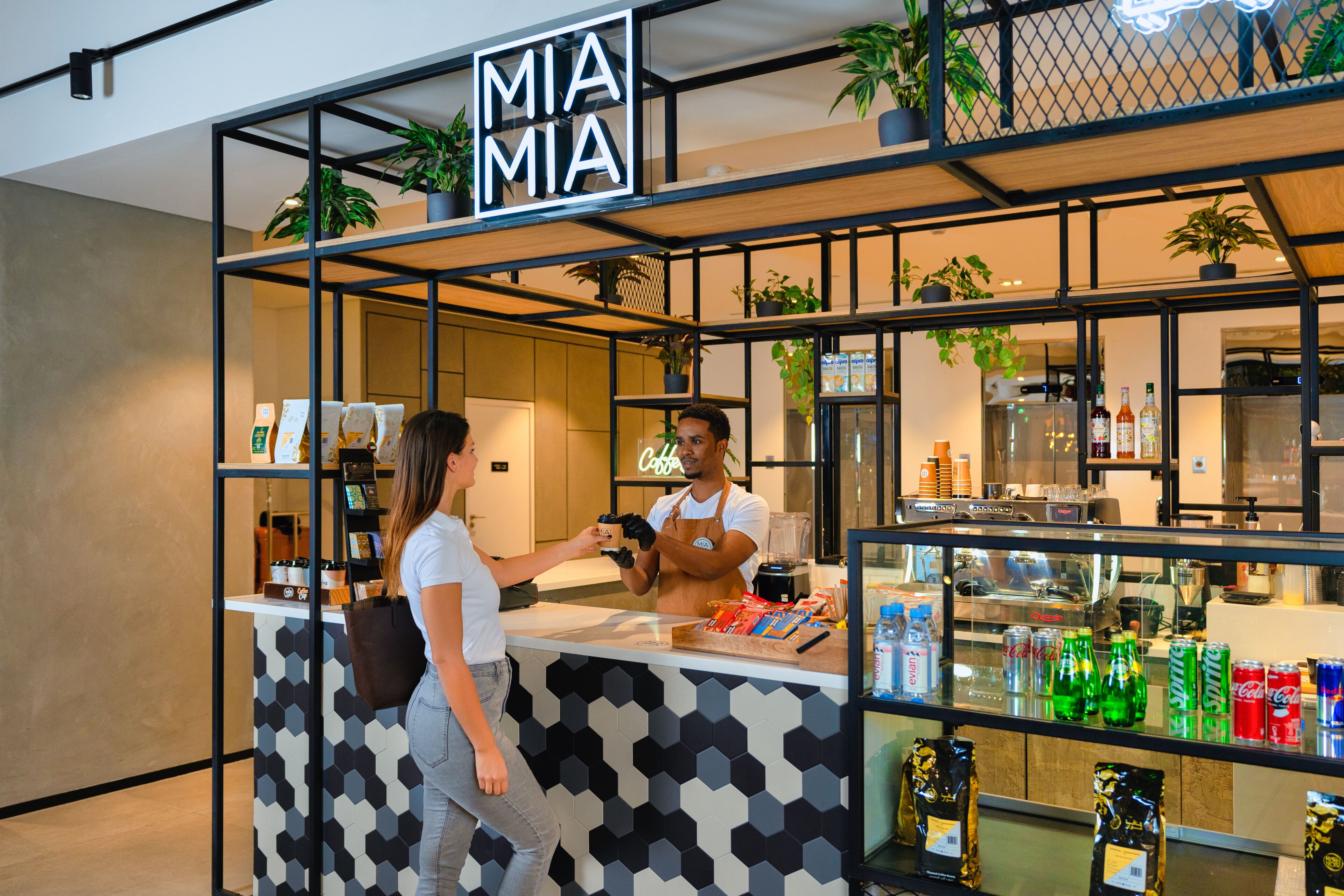 A coffee shop with a modern design, featuring a counter with a geometric pattern, a variety of beverages, and a neon sign.