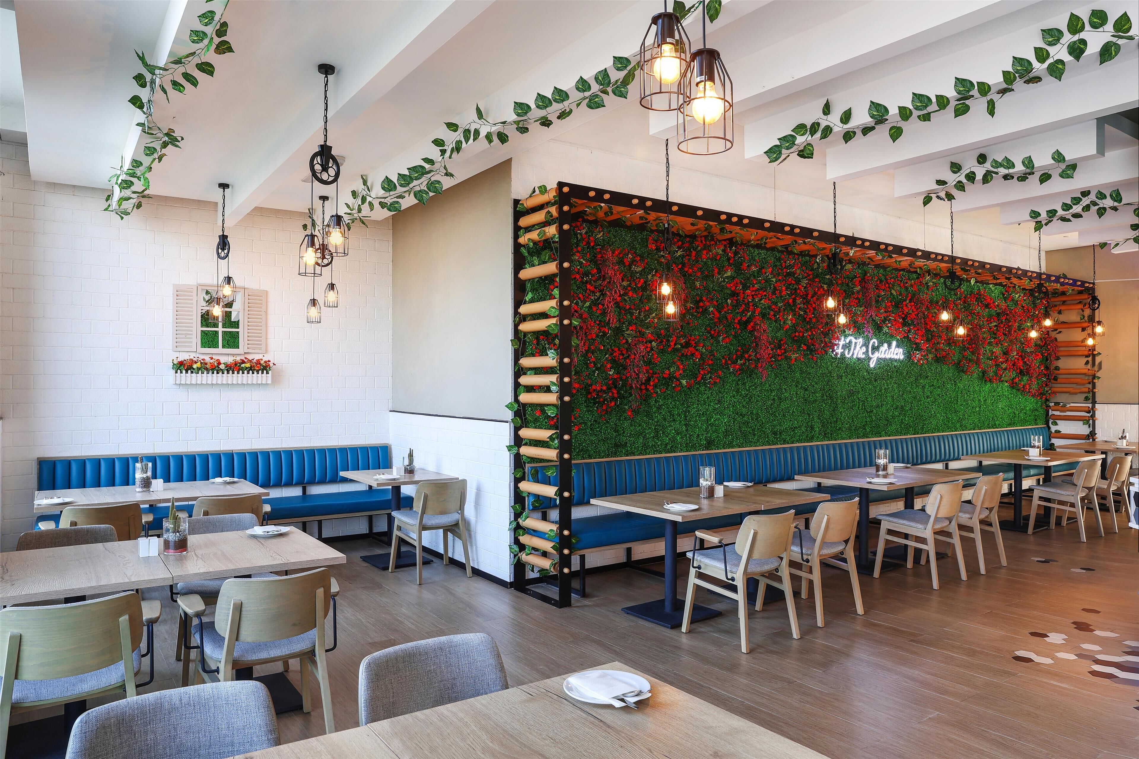 Een restaurant met houten tafels en blauwe bankjes, een groene wand met rode bloemen en hangende lampen.