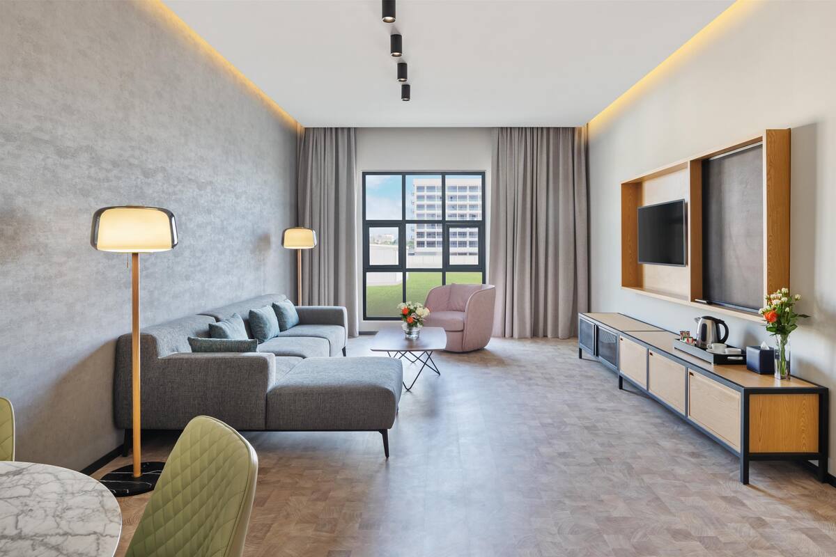 A modern hotel room with a grey sofa, a pink armchair, a dining table with chairs, a TV mounted on the wall, and a large window with curtains.