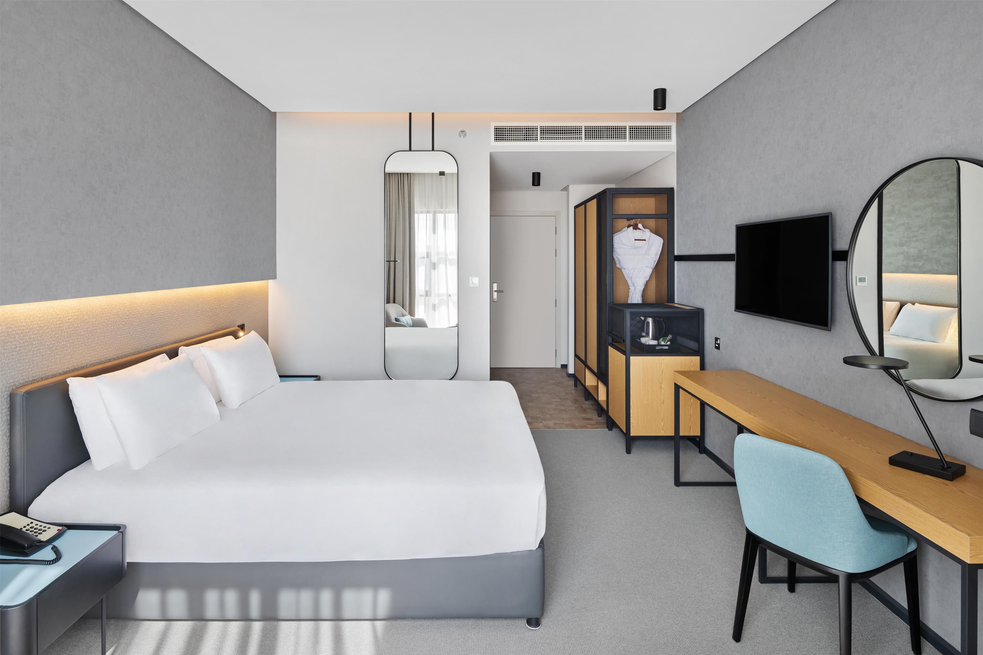 A modern hotel room with a large bed, a desk, a chair, a TV, and a wardrobe.