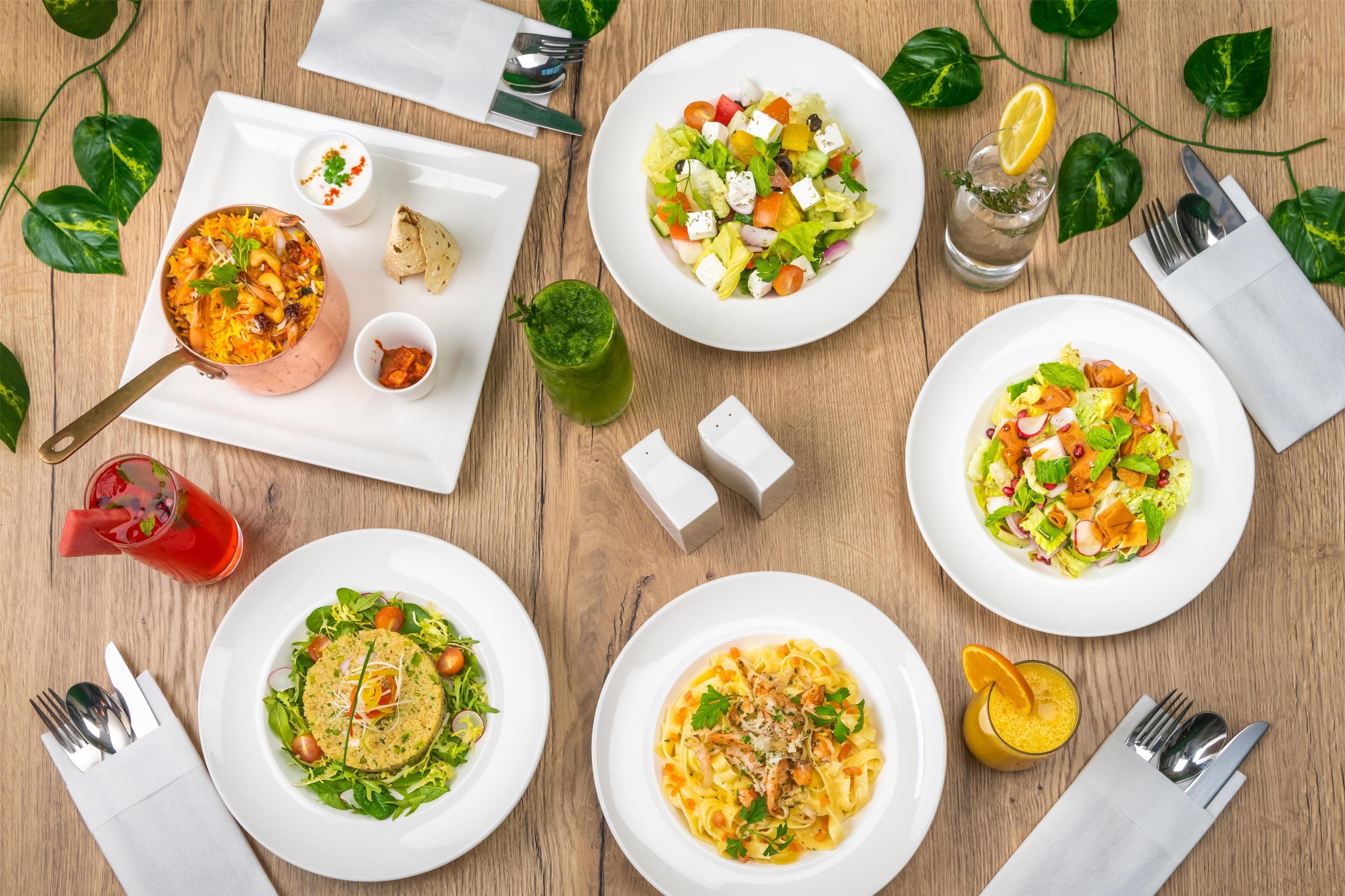 A wooden table with various dishes, including a bowl of rice, a salad, and pasta, along with condiments and drinks.