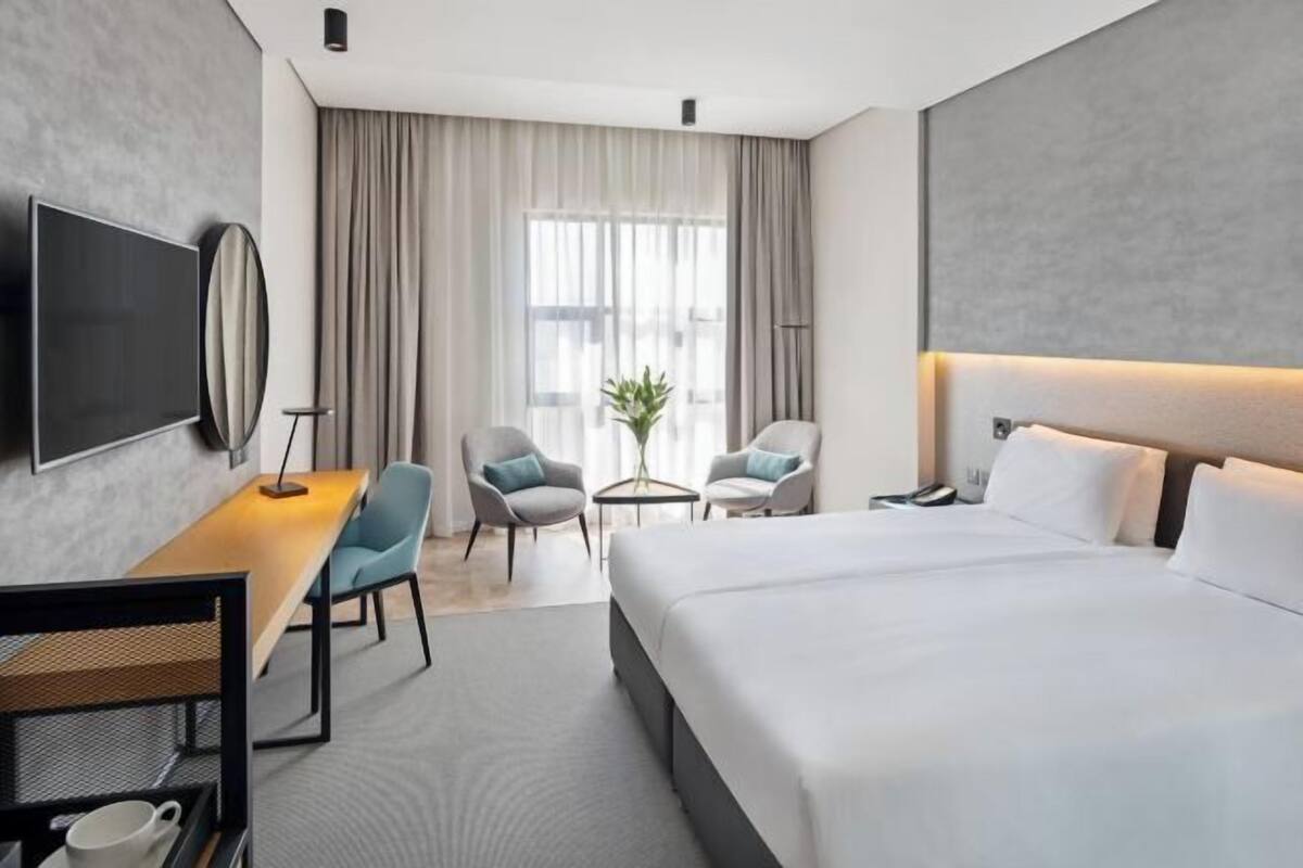 A modern hotel room with a large bed, a desk with a chair, a small table with a plant, and a TV mounted on the wall.