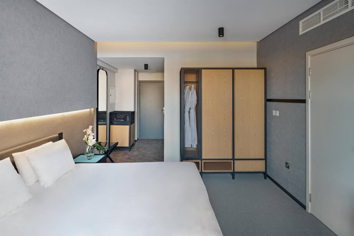A modern hotel room with a large bed, a bedside table with a vase of flowers, a walk-in closet, and a bathroom visible through an open door.