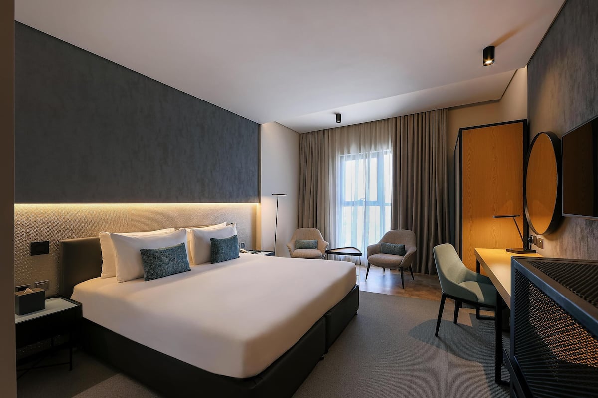 A modern hotel room with a large bed, a desk, two chairs, a flat-screen TV, and a large window with curtains.