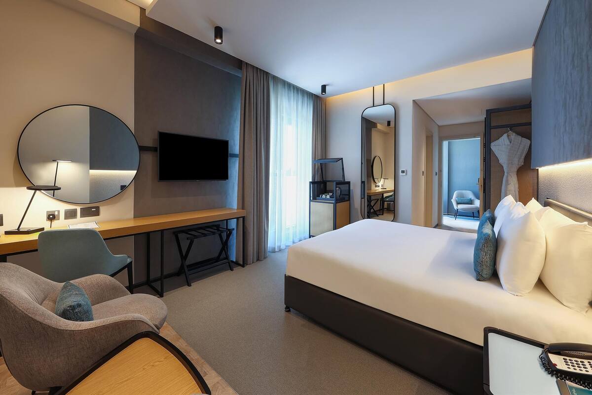 A modern hotel room with a large bed, a desk with a chair, a television, and a mirror.