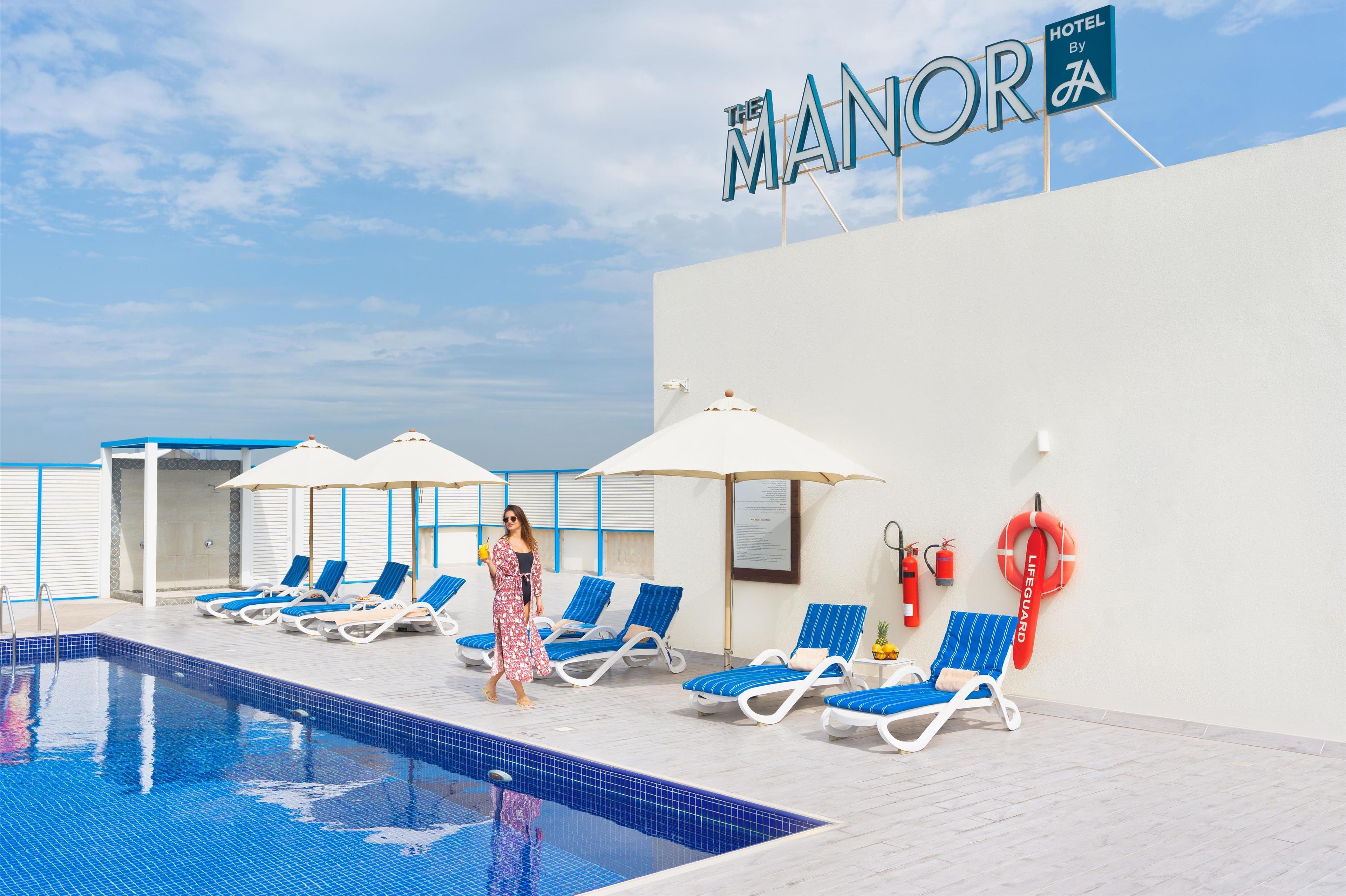 Zona de piscina de un hotel con sillones de descanso y sombrillas, un puesto de socorrista y un cartel que dice "The Manor Hotel".