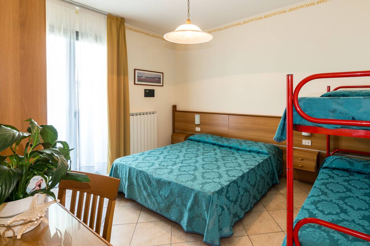 Une chambre avec deux lits, un bureau en bois, une chaise, une plante en pot et une fenêtre avec des rideaux.