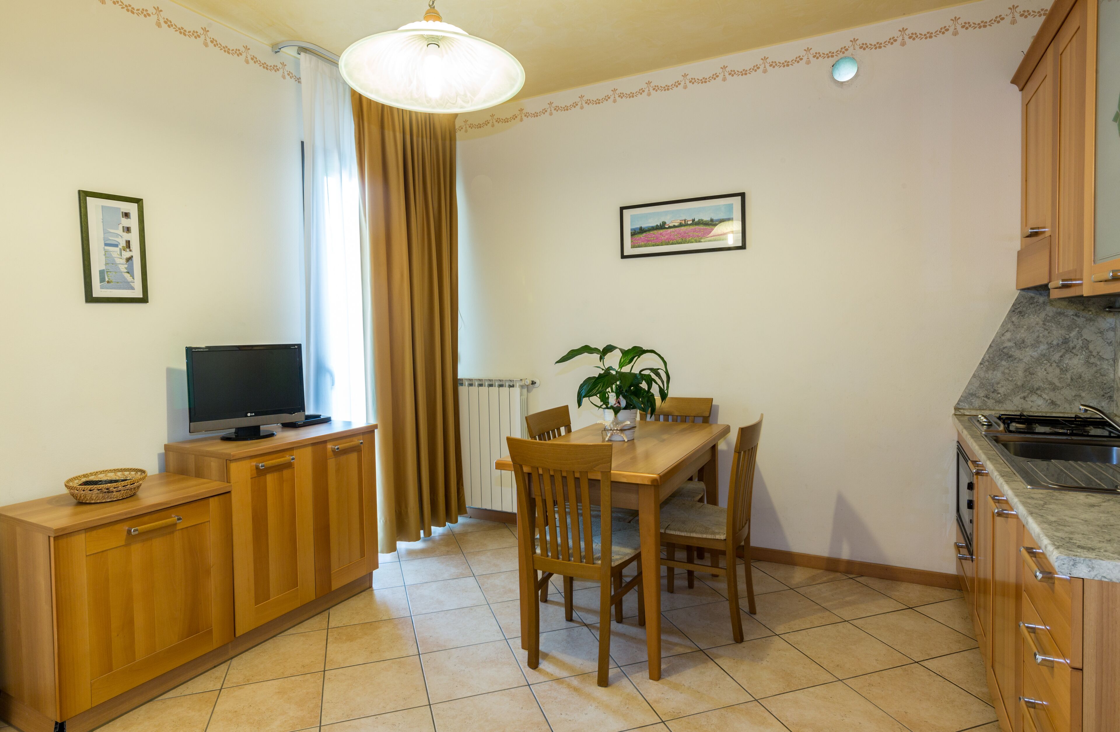 A compact kitchen with wooden cabinets, a dining table with chairs, a television on a cabinet, and a potted plant.